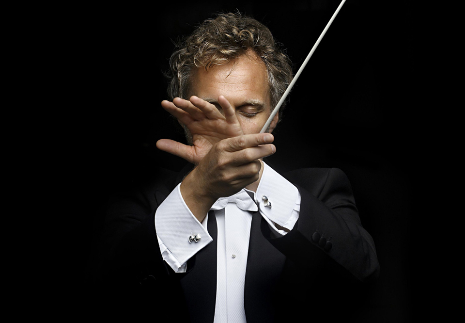 Thomas Søndergård with both hands in front, partly covering his face, holding the baton and making a serene conductor's pose. Photography.