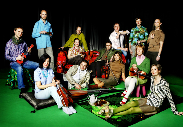 Gruppbild av tretton musiker, där vissa står och vissa sitter på golvet. Färglada kläder och miljö. Rebaroque. Fotografi.