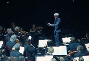 Man som dirigerar stor orkester i ljussatt blått sken. Utdrag ur filmen.