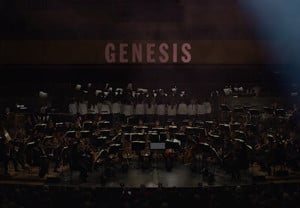 Utdrag ur filmen Genesis. Stor orkester och upplyst i bakgrunden med tetxen Genesis. Fotografi.