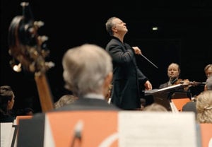 Dirigent med bakåtlutat huvud titar ut över stor orkester. Utdrag ur filmen.