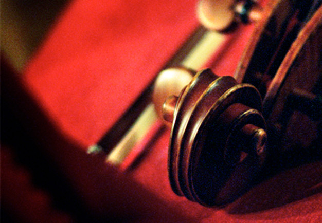Närbild på snäckan på en cello lutandes mot en stol med röd dyna. Fotografi.