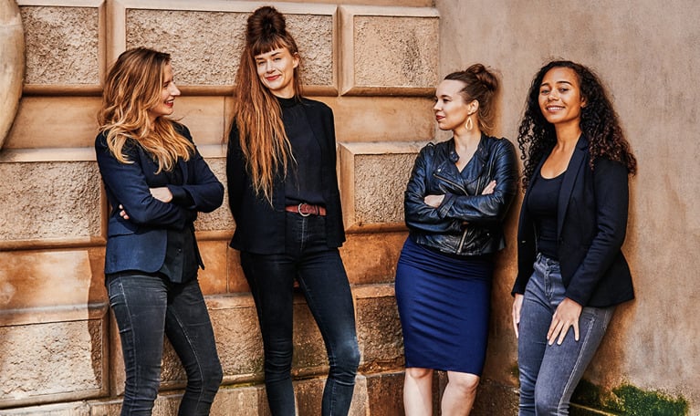 Four women leaning against a wall. Photo.