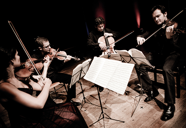 The quartet is sitting down playing in a dimly lit environment. Photography.