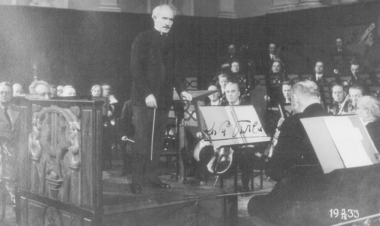 Stående, svartklädd man tillsammans med sittande musiker. Foto av den italienske dirigenten Arturo Toscanini på podiet med orkestern i Konserthuset 1933.