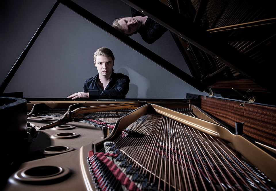 Koncentrerad pianist som sitter bakom flygeln. Locket är uppfällt på flygeln så man ser tangenterna. Denis Kozhukhin. Fotografi.