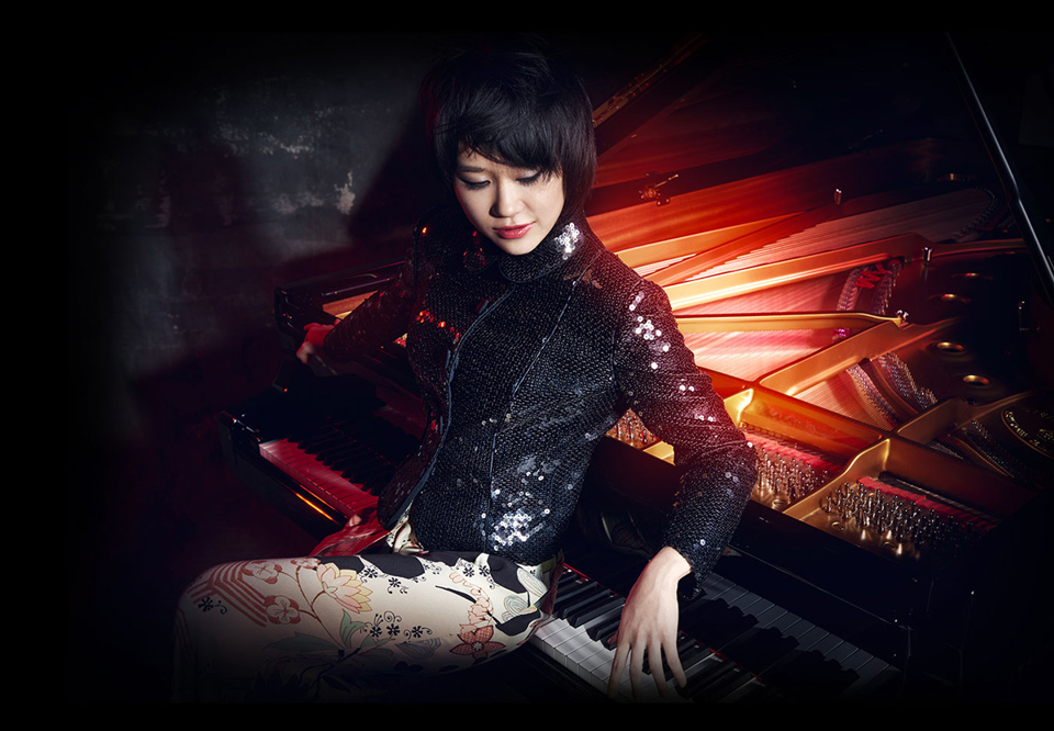 Pianist sittandes vid flygel med blommiga byxor och svart topp. Yuja Wang. Fotografi.