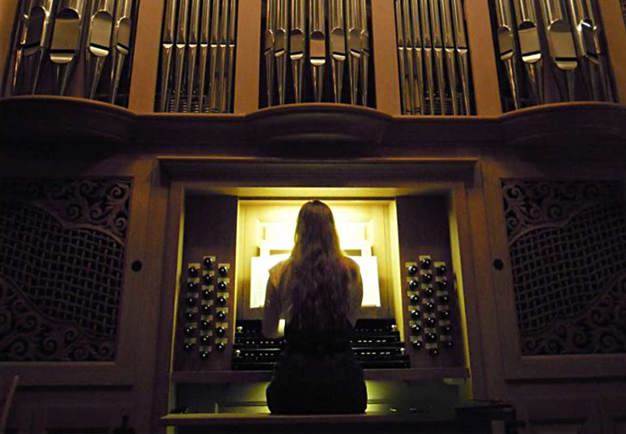 Kvinna med långt hår sitter med ryggen mot kameran. Spelar på en magnifik orgel. Fotografi.