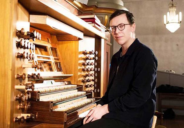 Photography of man in black suit and dark glasses. Sitting down in front of the organ and looking into the camera. Photography