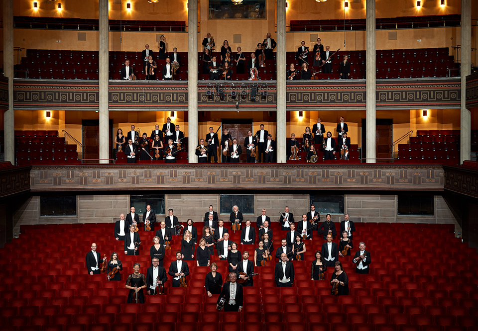 Musiker som står upp bland stolarna i Stora salen. Fotografi.