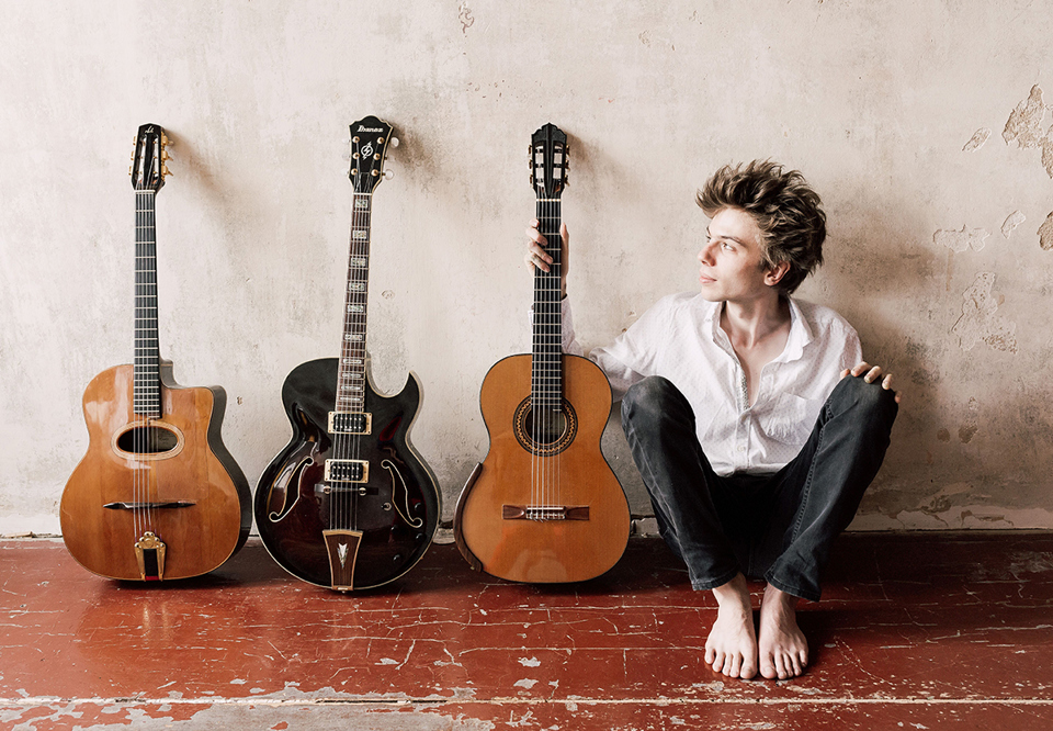 Ung kille som sitter på golvet med tre gitarrer bredvid sig. Fotografi.