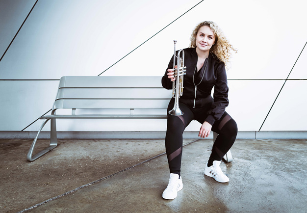 Kvinna som sitter på en bänk med trumpet i handen. Fotografi.