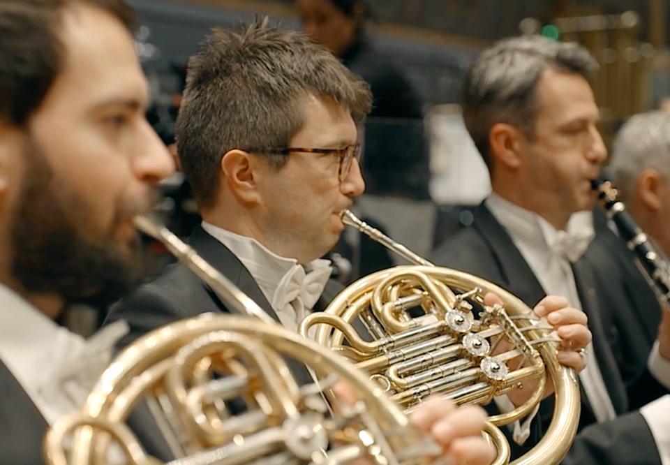 Närbild på man som spelar horn, i bakgrunden fler musiker. Konsertklädda. Fotografi.