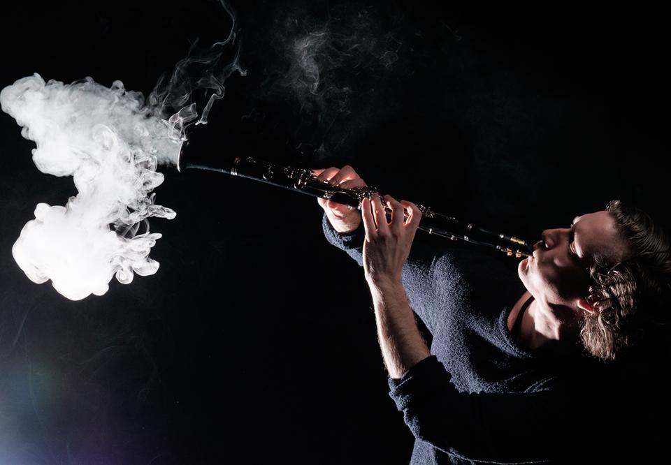 Man plays on a clarinet, smoke goes out from the bottom of the instrument. Dramatic photography.