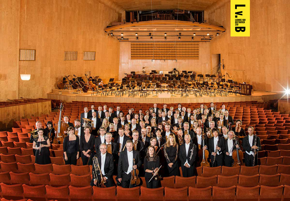 Gruppbild på frackklädda musiker som står bland röda stolar i en sal.Foto