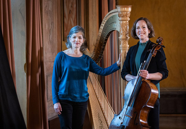 Två kvinnliga musiker, en står i förgrunden och håller en cello, den andra står lutad mot ett piano.