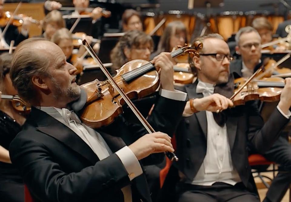 Konsertklädda musiker på scen, i närbild en man som spelar på sin fiol. Fotografi.