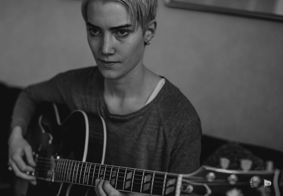 Svartvitt fotografi av en kvinna som sitter i en soffa och spelar gitarr.