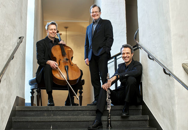 Fotografi av tre män, en sittande med en cello, en sittandes med en klarinett och en som står. Kostymklädda.