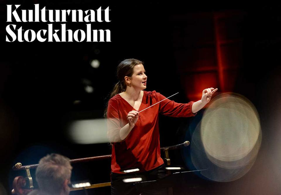 Kvinna i röd överdel med taktpinne dirigerar. Fotografi. 