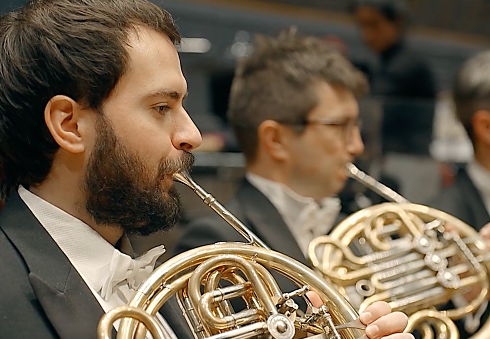 Närbild på man som spelar horn, i bakgrunden fler musiker. Konsertklädda. Fotografi.