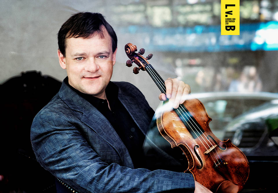Sittande man i mörk skjorta och kavaj, med violin i handen . Fotografi.