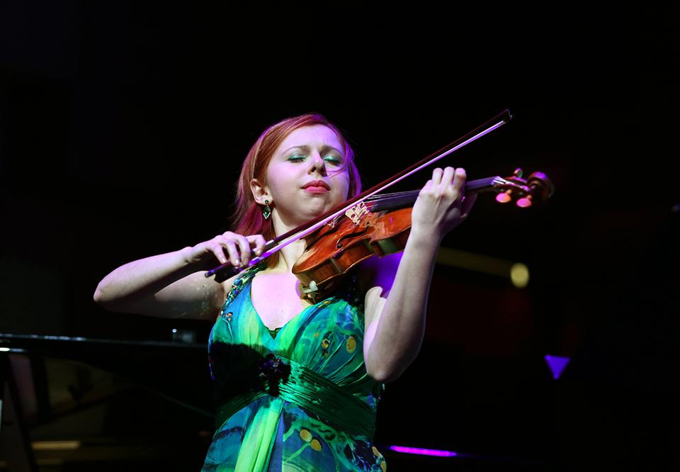 Fotografi av en kvinnlig violinist i ljust hår och grönt smink. Effektfullt ljussatt
