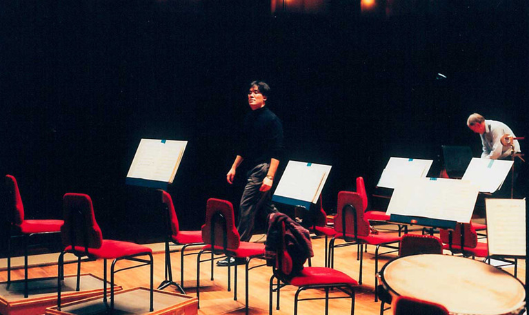 Tom scen med notställ och röda stolar. Svartklädd man längst bort i bild går över scenen. Fotografi.