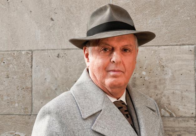 Dirigent lutandes mot grå vagg med ytterrock och hatt. Daniel Barenboim. Fotografi.