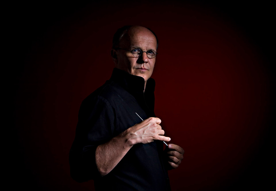 Man mot röd bakgrudn med dirigentpinne i handen. Fotografi.