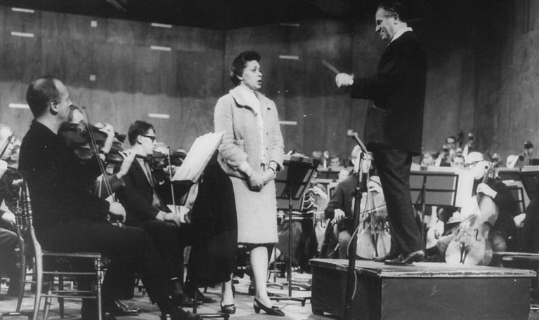 Kvinnlig sångare och manlig dirigent och delar av en orkester, sedda snett framifrån. Äldre gruppfotografi. Elisabeth Söderström med Antal Dorati och orkestern, vid Paris-gästspelet i Salle Pleyel i oktober 1966.
