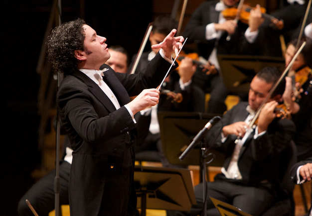 Gustavo Dudamel dirigerar, står på scen med musiker i bakgrunden. Fotografi.