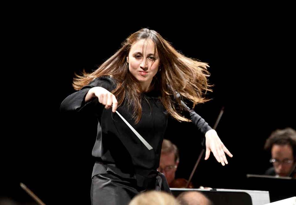 Kvinnlig dirigent på podiet i svart klädsel. Fotografi.