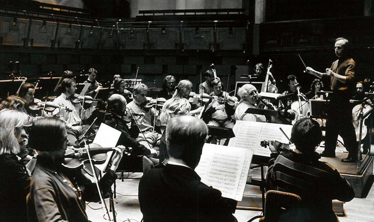 Black and white photograph from a rehearsal in the main hall, the conductor is viewed from the side as well as large parts of the musicians.