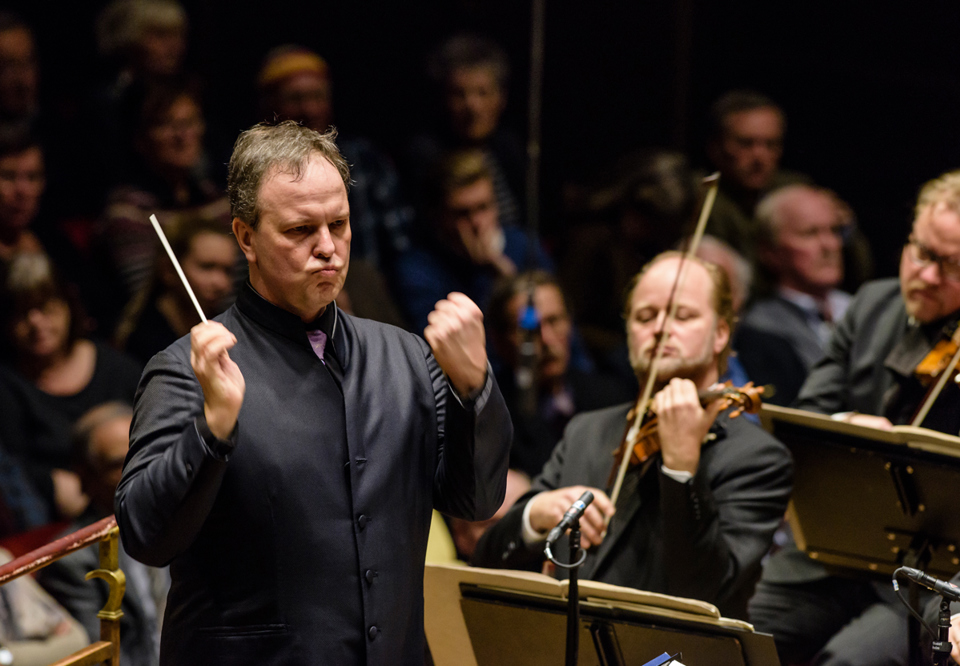 Intense moment of Sakari Oramo conducting the Royal Stockholm Philharmonic Orchestra, both hands raised and a grim expression on his face. Photography.