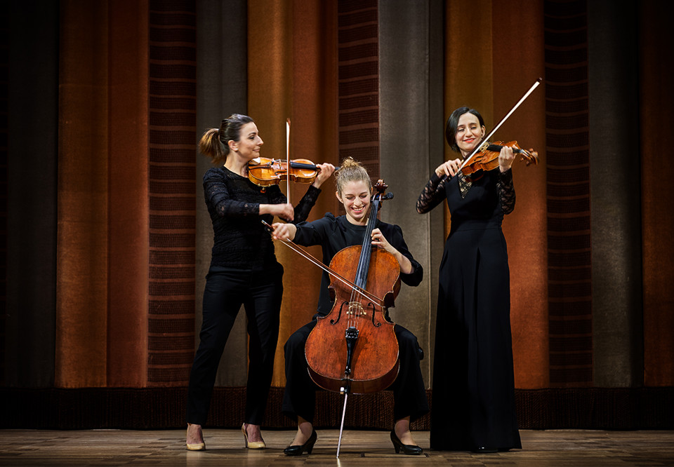 Three women on stage. Two plays the violin and one plays cello. Photo.