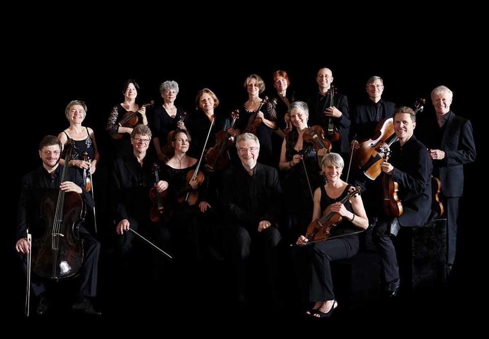 Freiburger Barockorchester är med i en gruppbild. Mörkbakrund, medlemmarna är festklädda och håller sina instrument. Ser glada ut. Fotografi.