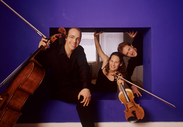 Three people with string instruments come out of a frame-like box.