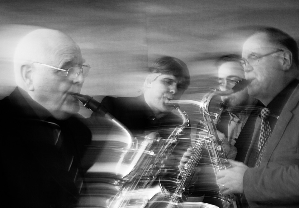Fyra personer som spelar saxofon, en svartvit bild där man fångar musikerna i en rörelse. Fotografi.