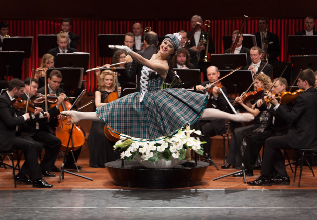 Orchestra playing in the background, a female ballet dancer making a grand jeté in front of them, smiling to the audiece. Photography.