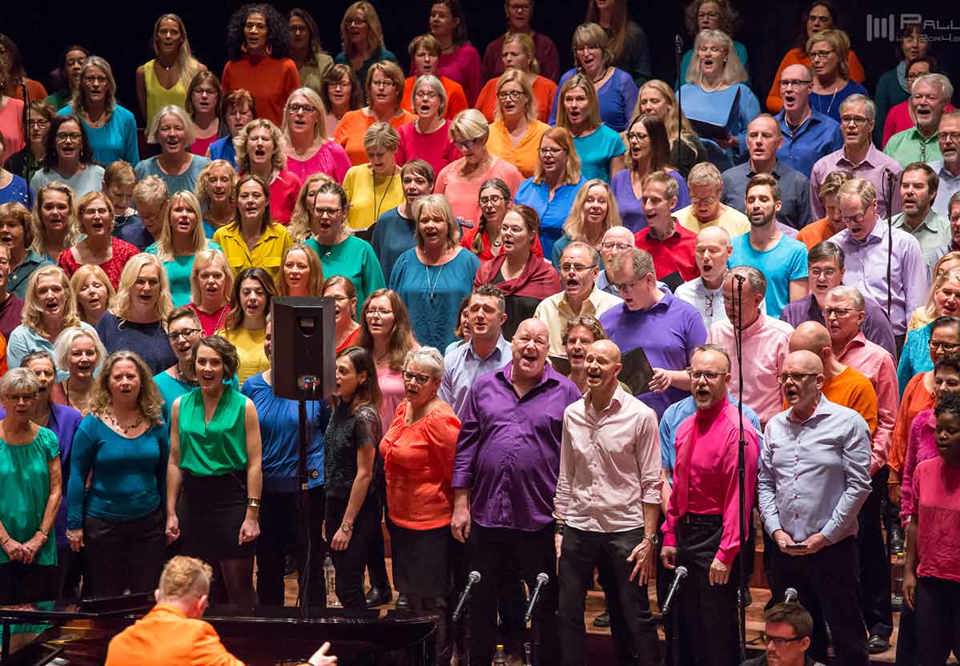 En närbild på en kör, många peraoner i olika kläder står och sjunger i färgglada kläder. Huller om buller. Foto.