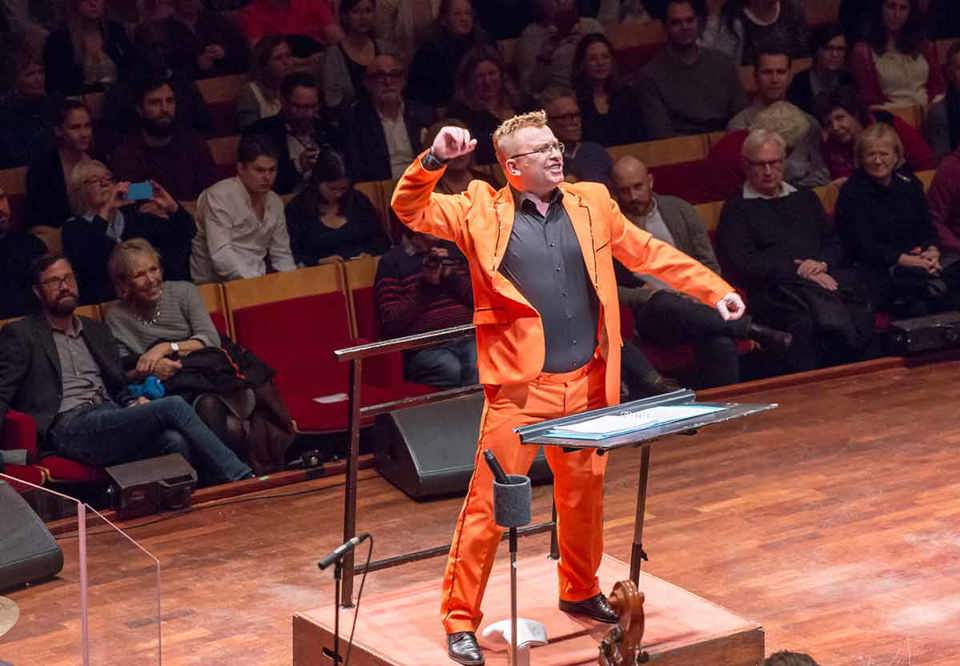Man i orange kostym står på scenen och sjunger. Fotografi från ett konserttillfälle.