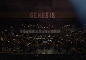 Utdrag ur filmen Genesis. Stor orkester och upplyst i bakgrunden med tetxen Genesis. Fotografi.