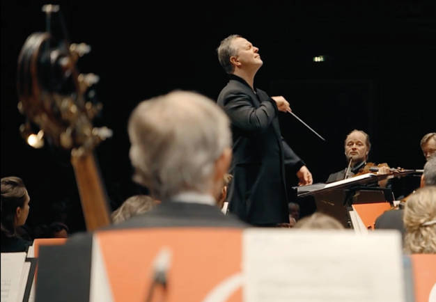 Dirigent med bakåtlutat huvud titar ut över stor orkester. Utdrag ur filmen.