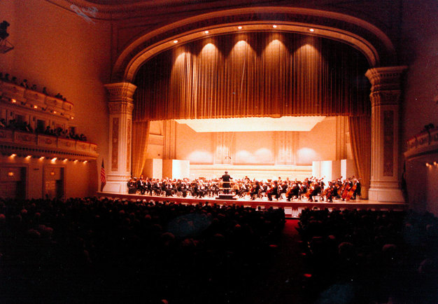 Upplyst scen sedd på avstånd från publikplats. Gruppfotografi av Yuri Ahronovitch och orkestern i Carnegie Hall den 31 oktober 1984.