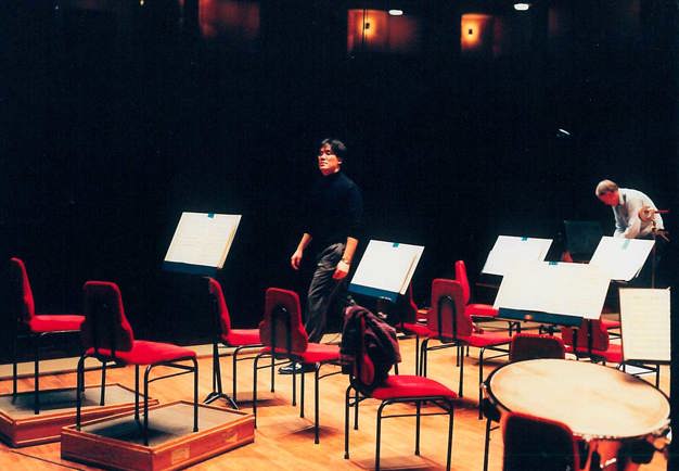 The conductor walking off the stage after a rehearsal, empty seats around him on the podium. Photograph.
