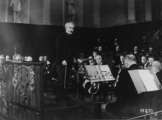 The conductor talking to the orchestra during rehearsal. Black and white photograph.