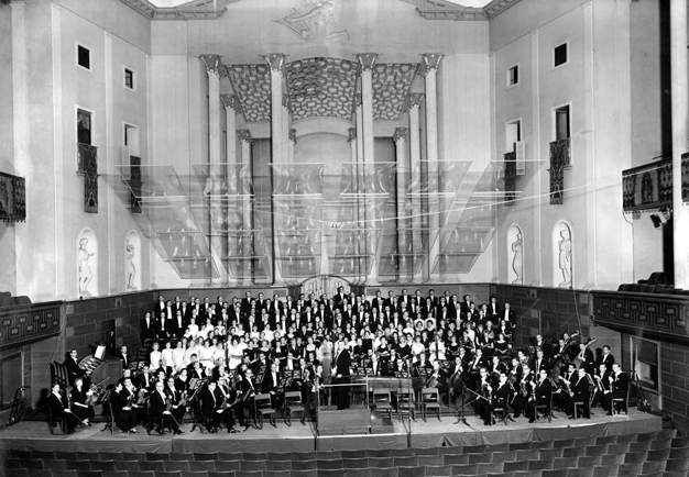 Black and white photo of the conductor and the orchestra posing in the main hall of Konserthuset.