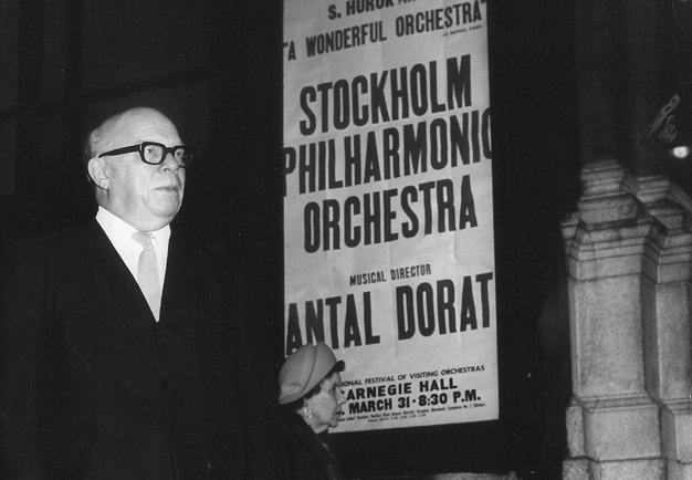 Black and white photograph of the director standing, posters of the orchestra's concert are clearly visible in the background.
