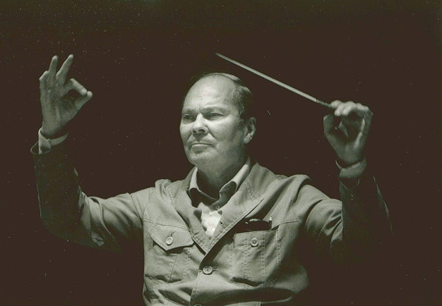 Man sedd framifrån, klädd i skjorta och med höjda armar, en dirigentpinne i ena handen. Den finländske dirigenten Paavo Berglund dirigerar. Svart-vitt fotografi.